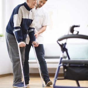 Elderly man walking on crutches and a helpful male nurse supporting him