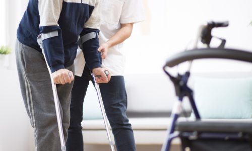 Elderly man walking on crutches and a helpful male nurse supporting him